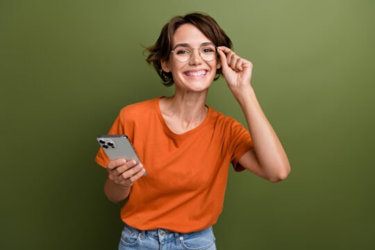 Portrait of smart girlish person wear stylish t-shirt touching eyewear holding smartphone read email isolated on green color background.